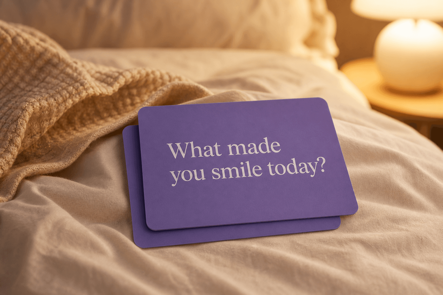 A stack of lavender question cards on a bed, the top card reading: What made you smile today?